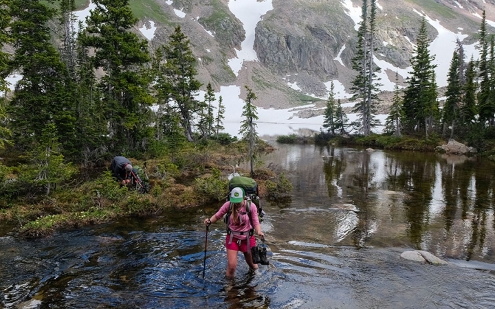 A person wearing a backpack uses a trekking pole as they walk through a shallow alpine lake.